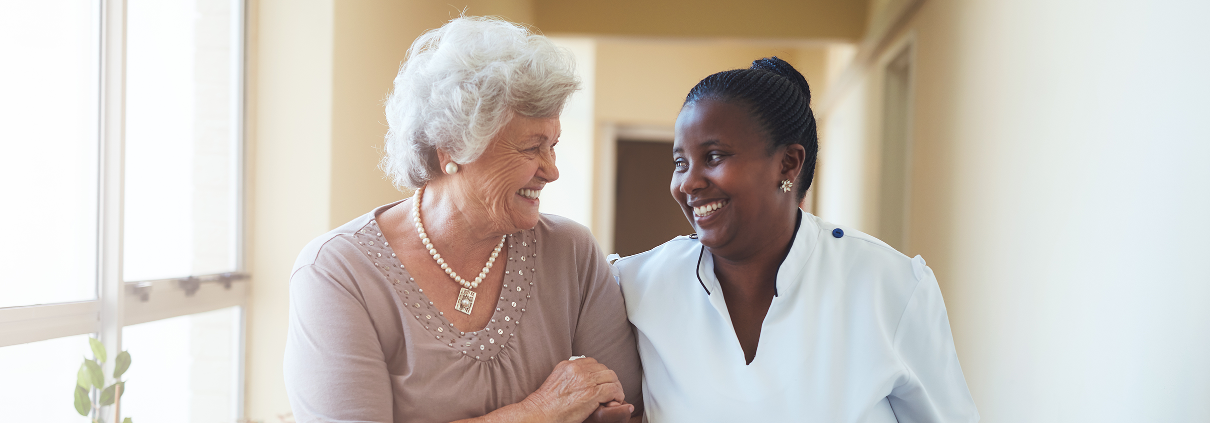 A senior woman with short white hair smiling and holding onto the arm of her female caregiver