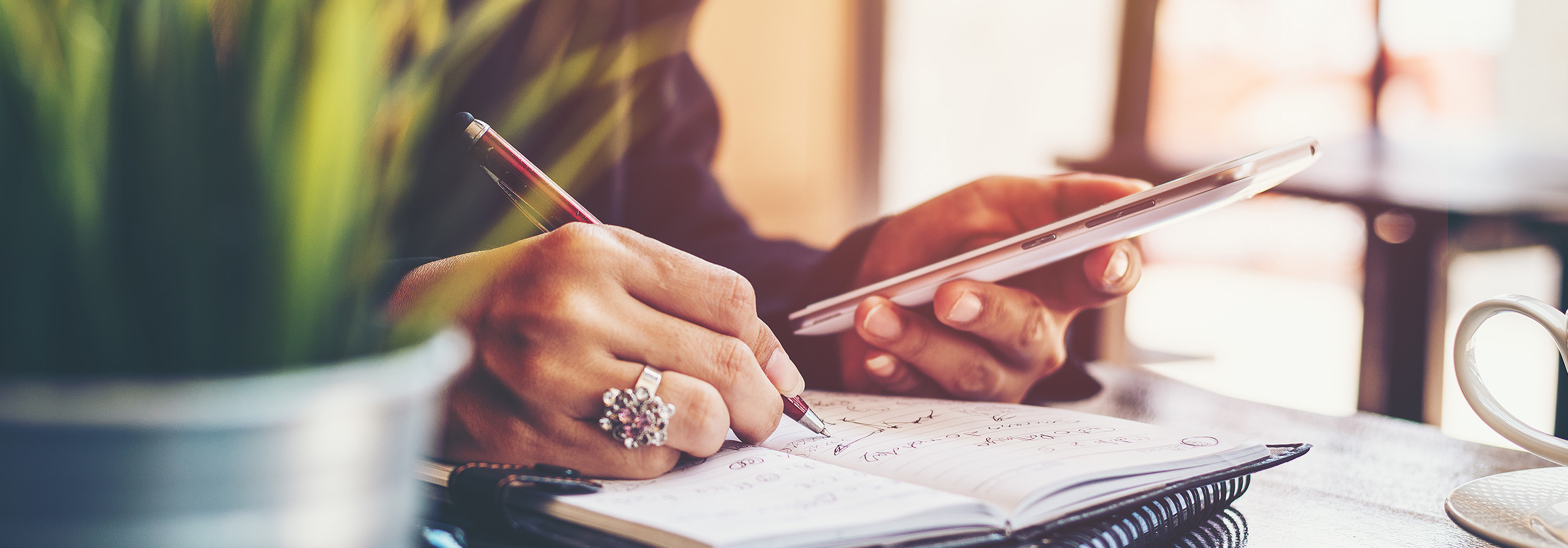 Close up of a person's hands as they use their right hand that has a flower ring on it to write in a notebook and they use their left hand to hold their smartphone