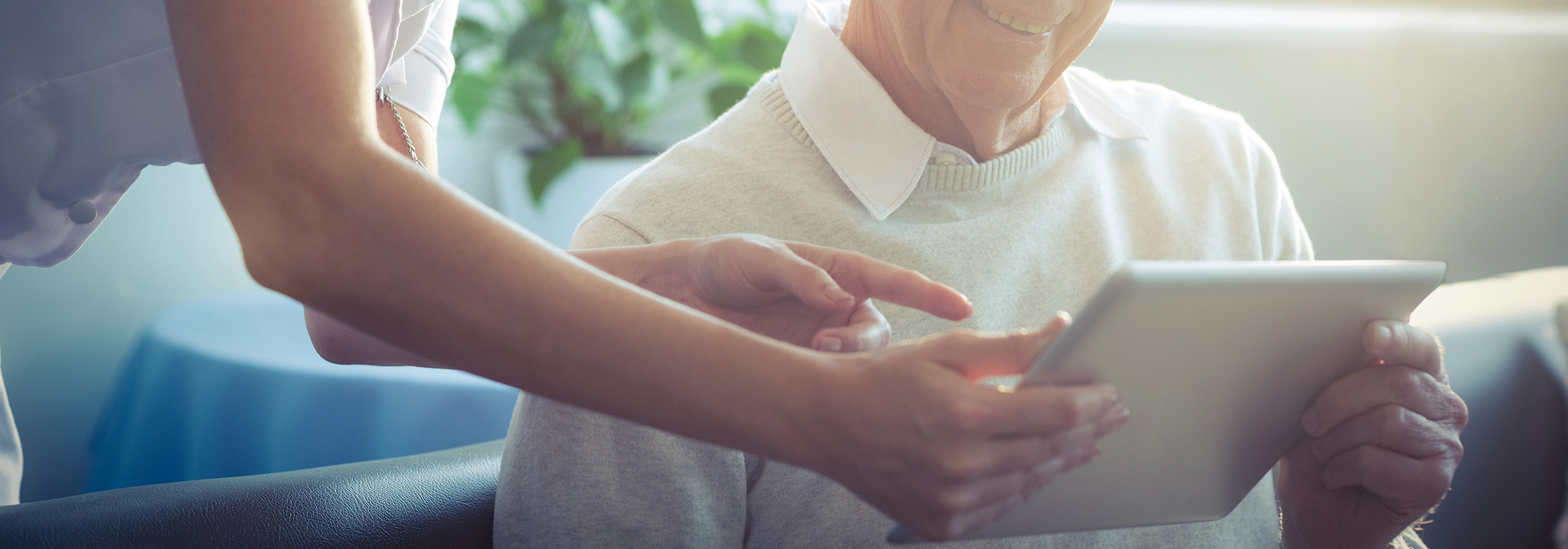 A senior man sitting on the couch smiling as a young woman hands an iPad to him