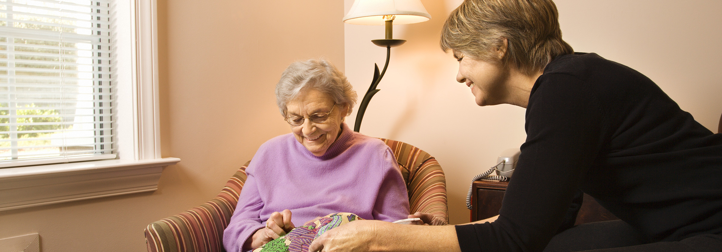 A senior woman wearing a purple sweater sitting down in a striped armchair sewing as her caregiver smiles and watches her