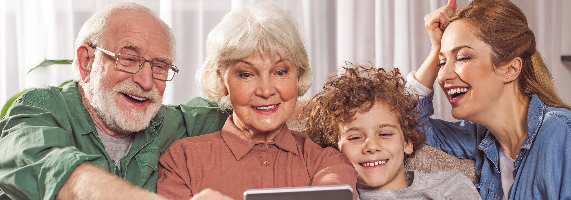 A senior man and woman sitting on the couch looking down at an iPad smiling and laughing with their daughter and grandson
