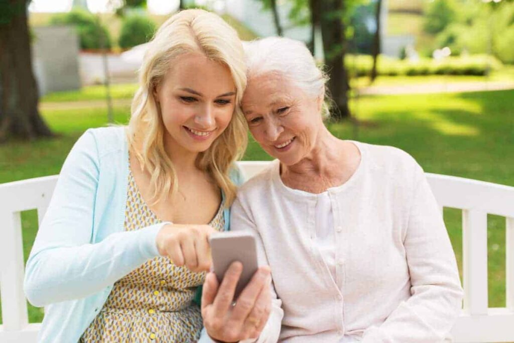 Senior woman sitting outdoors on a bench with her daughter looking at information for Traditions at Bridgepointe Gardens on a smartphone