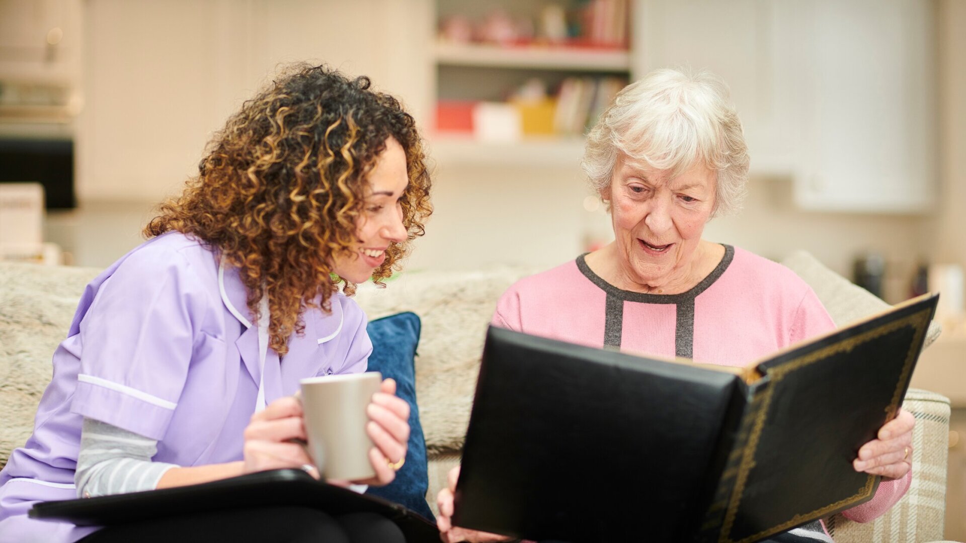 Female memory care resident sitting on a couch in her memory care apartment at Traditions at Bridgepointe Gardens with a caregiver