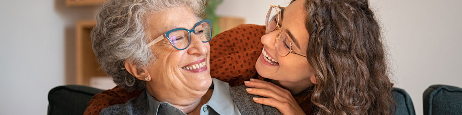 A senior woman wearing blue glasses smiling and looking at her daughter who has her arms around her shoulders