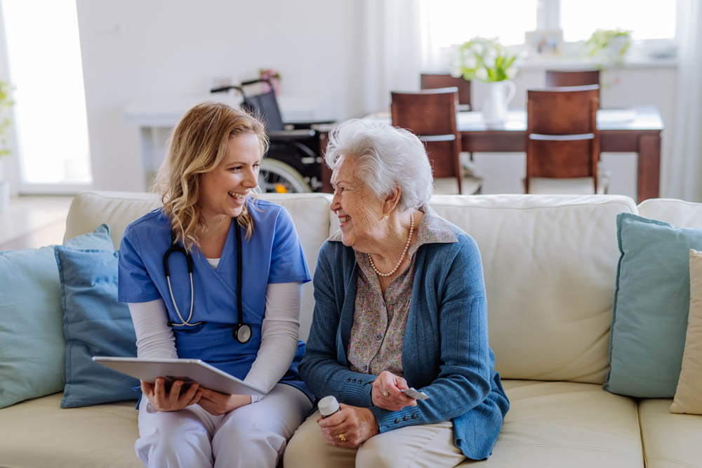 Female nurse sitting on a sofa with a memory care patient, laughing and conversing, at Traditions at Bridgepointe Gardens in Jeffersonville, IN.
