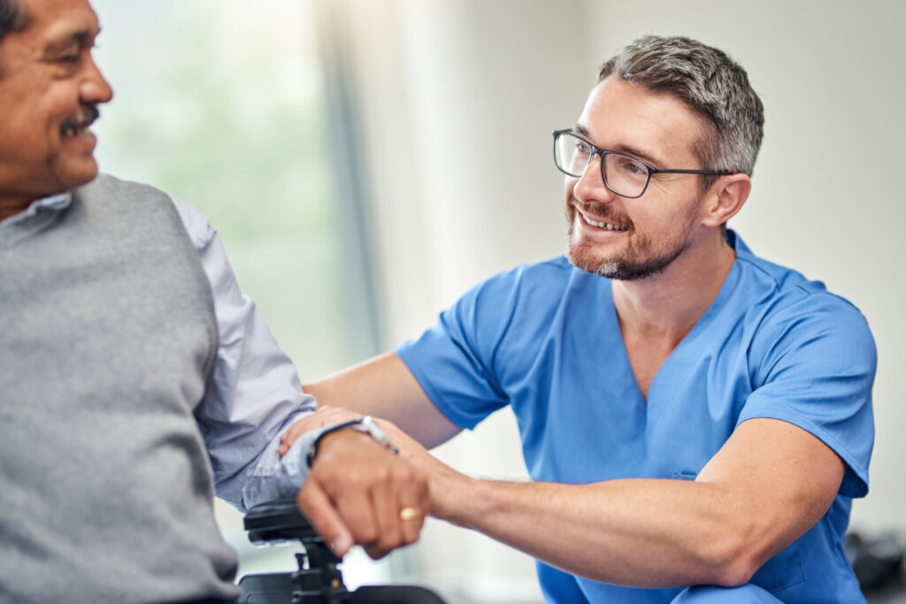 Male nurse smiling and patting the arm of a memory care resident at Traditions at Bridgepointe Gardens in Jeffersonville, IN.