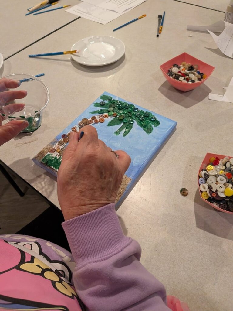 A resident carefully places decorative buttons onto a painted canvas palm tree scene during a mixed-media art activity at Traditions at Bridgepoint Gardens.