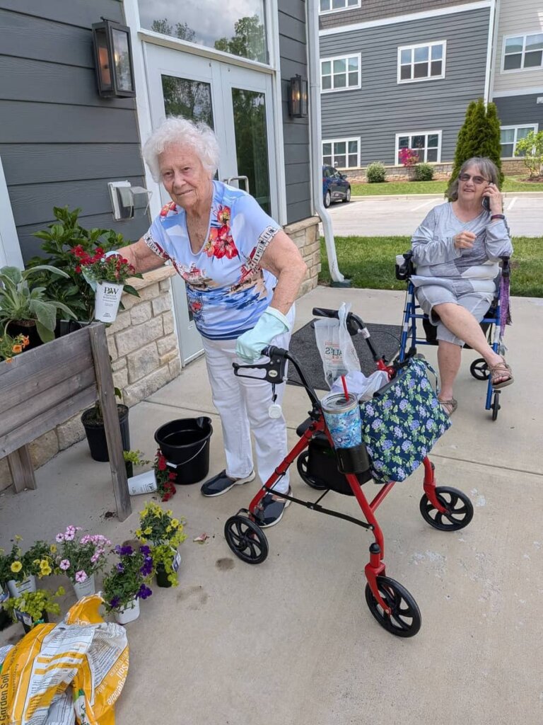 Two residents tend to a raised planter box filled with colorful flowers outside the Traditions at Bridgepoint Gardens community building on a summer evening.