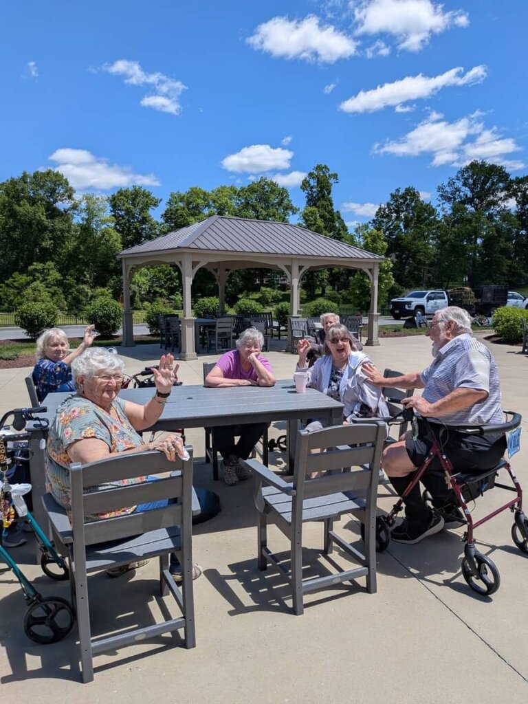 A group of residents waves and smiles at the camera while gathered around an outdoor patio table on a sunny day, with the community gazebo visible in the background at Traditions at Bridgepoint Gardens.