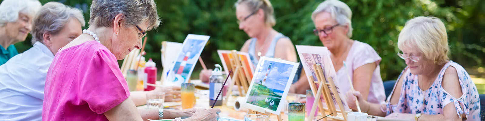 A group of senior women sitting outdoors at a table with a table cloth covering it painting during an event